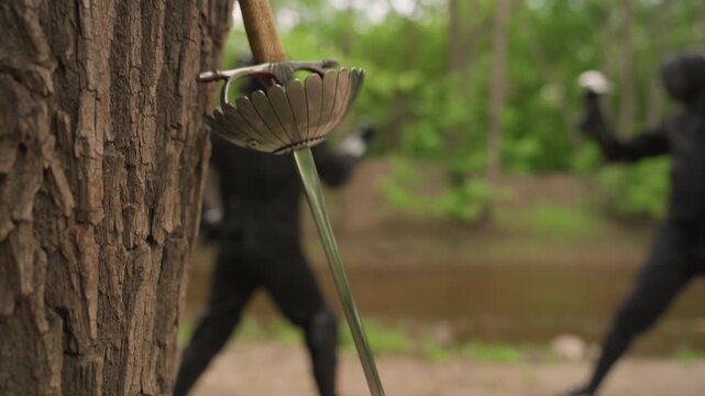 Close up of sword with wooden grip and metal basket hilt against tree bark while two people in black fencing gear spar outdoors. scene features natural green foliage background soft daylight