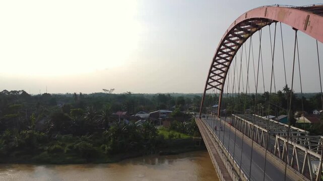 Aerial view of the red steel arch Wampu River Bridge on the Binjai - Langsa Toll Road spanning the river with lush tropical vegetation in Stabat, North Sumatra, Indonesia.