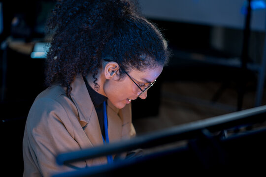 Black female data analyst cross-references code on a monitor with notes on a tablet in a blue-lit nighttime operations center.