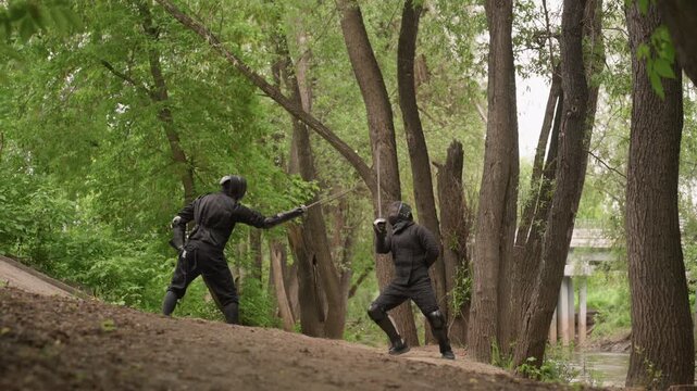 Two men in black ninja attire engage in sword fight on outdoor path surrounded by dense green trees. Their movements are fluid and focused with swords extended as they face each other