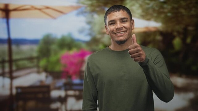 Man smiling and showing thumb while pointing finger toward camera in studio wearing green long sleeve shirt and goatee, casual pose conveys warmth; friendly approval.