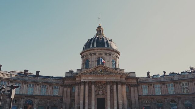 Static wide angle frontal shot of the majestic Institut de France facade featuring its iconic dome, classical columns, and French flag during warm sunset lighting in Paris. High quality 4K video.