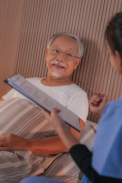 A female nurse in blue scrubs documents nursing care in pen while her senior patient dozes against his pillows, a real world scene of senior healthcare in Asia.