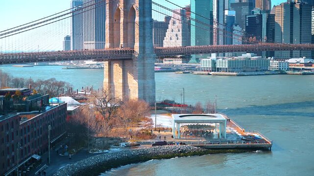 Brooklyn Bridge tower overlooking Dumbo waterfront. Snow patches dot the waterfront park as ferries pass beneath the stone bridge tower and Manhattan skyline.