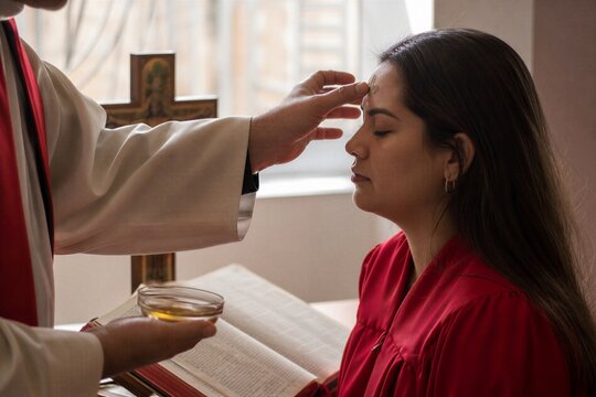 Priest anointing forehead of young woman with oil during christian confirmation ceremony in church. Sacrament of chrismation with cross sign and holy bible.