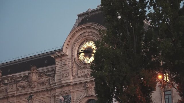 Moving shot from a Seine river cruise boat revealing the glowing giant station clock on the historic facade of Musee d Orsay behind tree foliage at dusk in Paris, France. High quality 4K B-roll video.