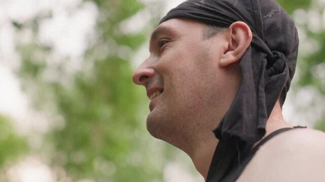 Close up of man wearing black tank top and head covering showing facial expressions in outdoor setting with greenery. subject skin glistens under natural light as camera maintains steady