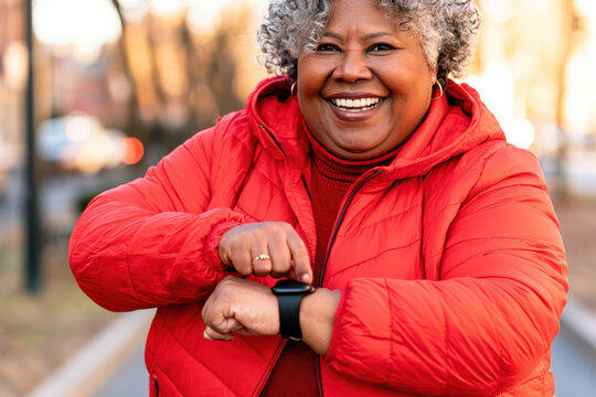 African American woman in red jacket smiles while checking smartwatch outdoors in a city park with trees and pathway visible in the background