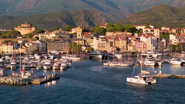Saint Florent Corsica 4K drone video. White catamaran sailing into luxury marina with telephoto 70mm lens perspective of coastal town. High end yachting travel b roll stock footage