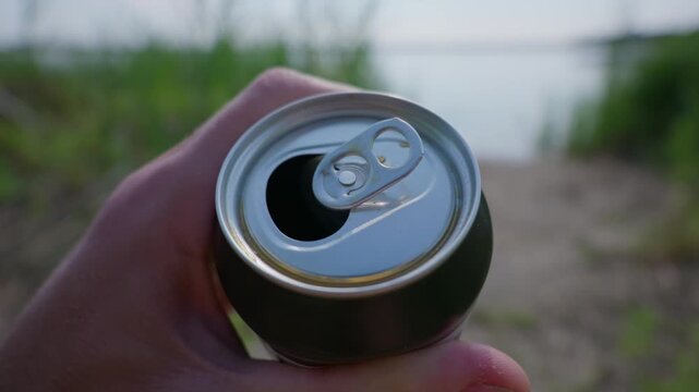 A hand holds an open aluminum beverage can with a metal pull tab in nature. Concept of outdoor beverage consumption, refreshing cold drinks, packaging design, and relaxation.
