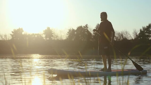 Supboard boad boy. A boy stands on a paddleboard, paddling gently on the water as the sun sets. The boy enjoys the peaceful scene, with reflections shimmering on the water.