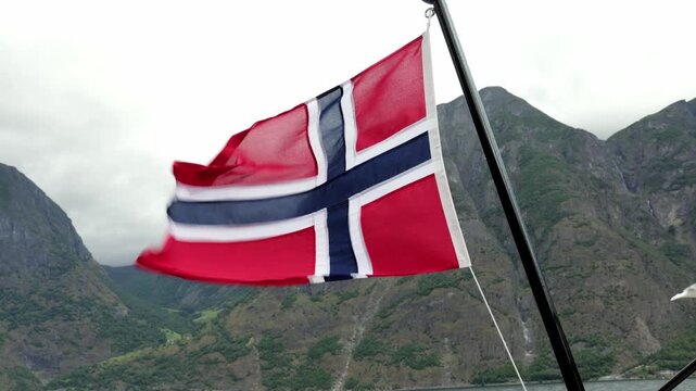 Norwegian flag waving in the wind against a backdrop of majestic mountains and cloudy skies, showcasing vibrant colors and dynamic movement in a natural setting