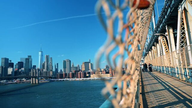 New York, USA, 1 April 2026 : Pedestrians pause on Manhattan Bridge overlooking skyline. Sunlight casts long shadows across the walkway as the Brooklyn Bridge spans the river beyond.