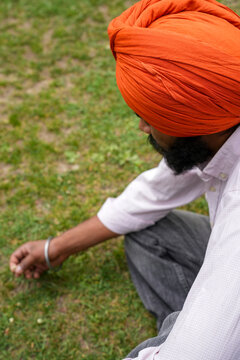 High angle close-up shot of a young Sikh man wearing a vibrant orange turban, sitting on lush green grass and carefully picking a tiny wildflower outdoors.