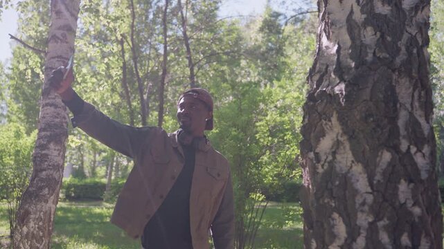 Close up of man taking selfie outdoors in park with birch trees and greenery. Man holds smartphone above head wearing brown jacket over black shirt headphones around neck. Bright daylight