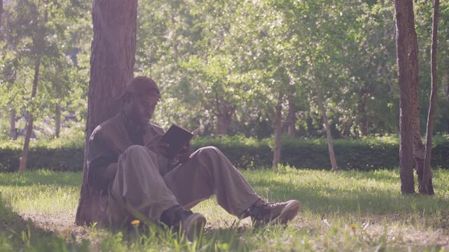 Man sits on grass under tree in outdoor park setting reading book. Man wears casual clothing including cargo pants and sneakers with sunlight filtering through trees creating soft shadows