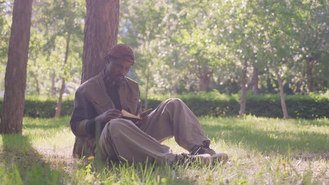Close up of man reading book outdoors under tree with headphones around neck. Soft natural light illuminates calm scene in park setting with green grass and trees background