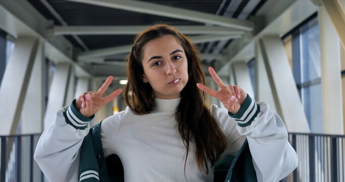 Young woman content creator posing, double peace signs, varsity jacket on walkway bridge with industrial beams, natural window light, playful confident expression, casual streetwear, social media