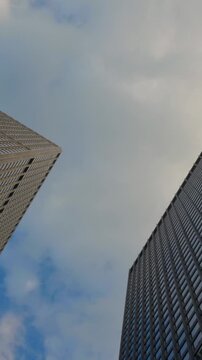 Skyscrapers rise against cloudy sky, upward view between building facades, glass tower and textured high-rise edges framing blue sky patches and drifting clouds within urban canyon scene