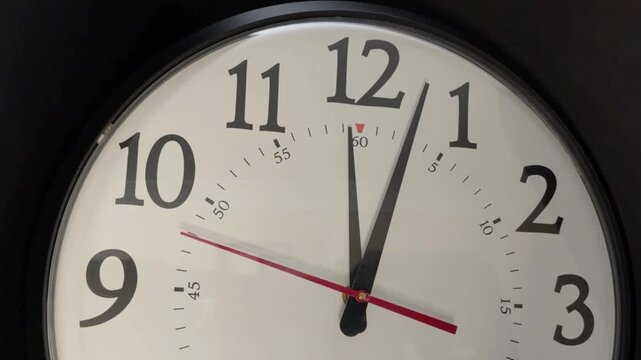 Frontal close-up of analog wall clock with red second hand near twelve, bold numerals across upper dial, glossy surface, precise office timepiece with cropped panoramic view