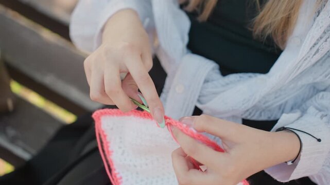 Close up of hands crocheting pink and white fabric with green hook showing detailed yarn work in progress. person wears light colored clothing while seated outdoors on wooden bench focused