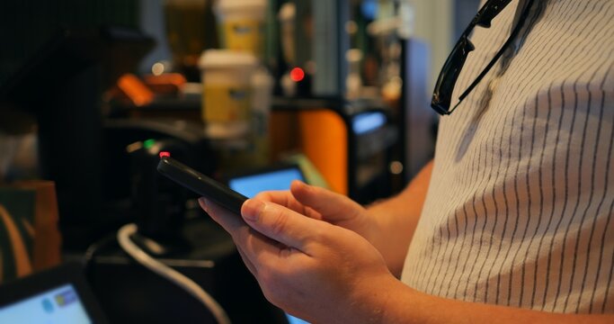 Person holding smartphone for mobile payment inside coffee shop, with blurred terminal screens and counter equipment in background, close view of hands and device, finger detail, contactless texture