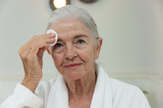 Senior female holding round cotton pad, cleansing temple at bathroom vanity wearing white bathrobe
