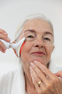 Senior woman holding triangular LED facial device on cheek at home showing gold ring and bathrobe