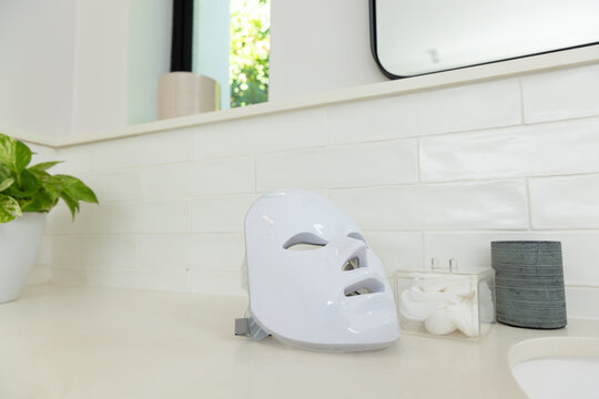 White LED facial mask resting on vanity countertop with cotton pads, planter, mirror, window