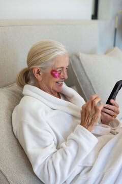 Senior woman reclining at home on couch in bathrobe, holding phone, showing pink heart eye patch