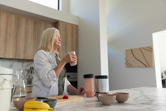 Senior woman standing in modern home kitchen holding pink smoothie near window with blender cups