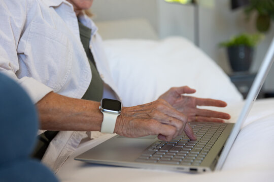Senior woman typing on silver laptop while wearing smartwatch with white band on white bedding