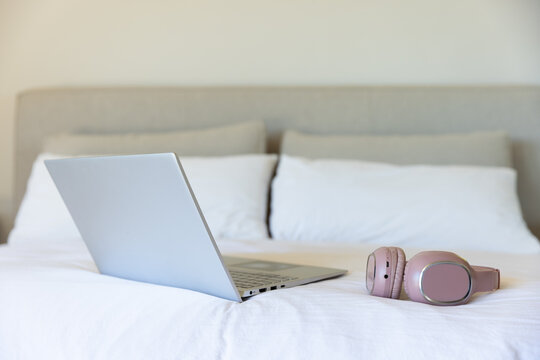 Silver laptop and pink headphones are resting on white bed in bedroom, soft natural light