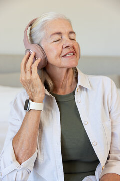 Senior female listening to music with pink headphones, wearing beige button-up shirt on sofa