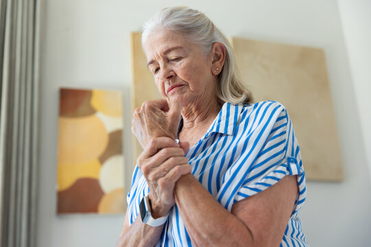 Senior woman standing at home holding left wrist while wearing smartwatch and striped shirt