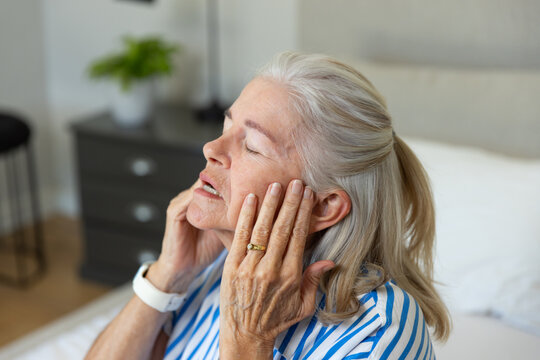 Senior woman sitting on bed pressing hands to temples in bedroom wearing striped top and wristwatch