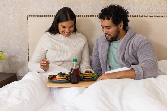 African couple in sweater and robe sitting in bed sharing tray with pancakes and green-capped syrup