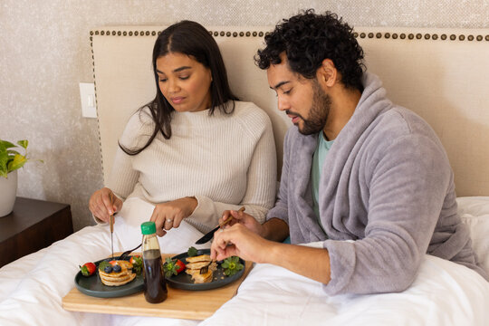 African American couple sitting bed sharing tray with pancakes, green syrup bottle, sweater, robe