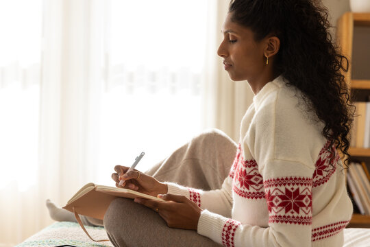 African woman writing in notebook cross-legged on bed wearing cream sweater red pattern