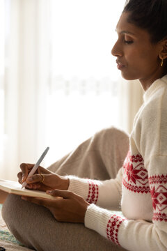 Asian adult female sitting near window in cream sweater writing on tablet with stylus, copy space