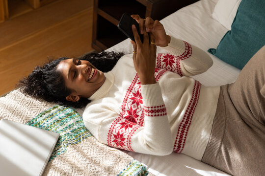 Woman lying on bed at home scrolling smartphone above face in white sweater laptop nearby