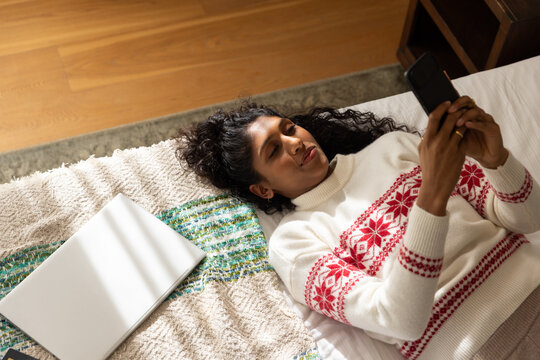 Woman lying on bed wearing white Nordic sweater, holding black smartphone, silver laptop nearby