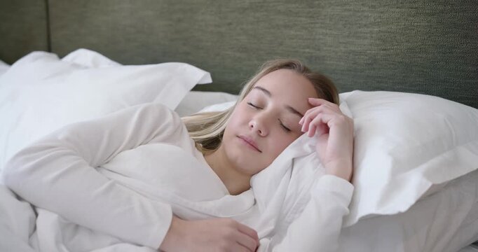 Soft window light lulling 20s woman lying on bed sleeping while breathing and shifting arm slightly