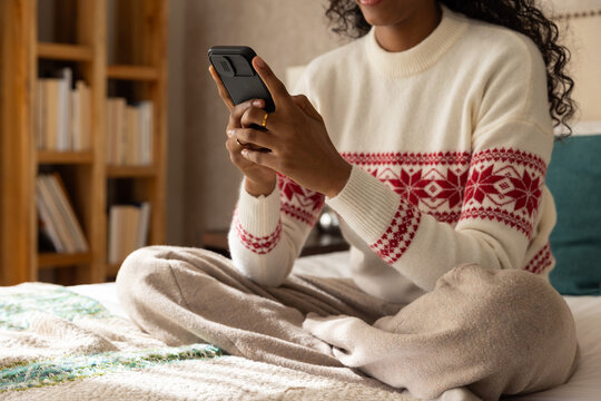 Woman sitting on bed holding smartphone and scrolling wearing knit sweater and showing gold ring
