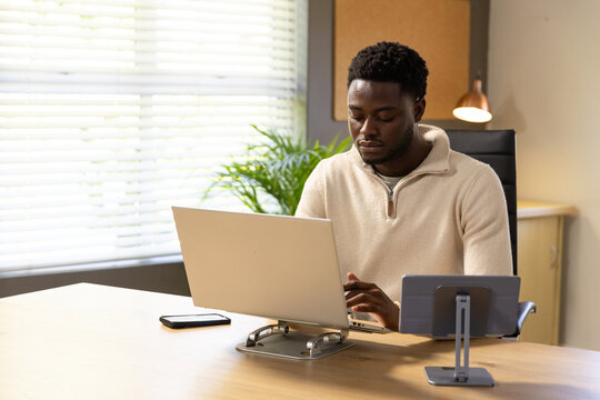 Man wearing beige pullover, sitting in office, working on laptop on stand with smartphone