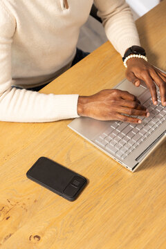 African American man typing on silver laptop at wood table, wearing cream pullover, watch, phone