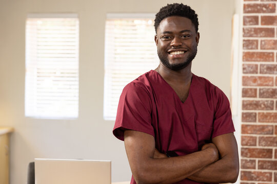 African adult male standing in maroon scrubs in office, smiling with closed silver laptop