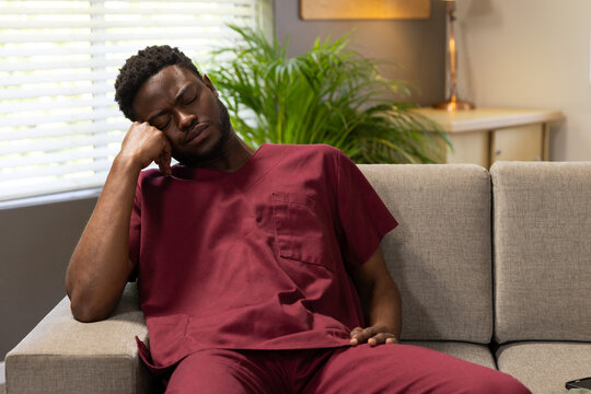 African male resting in lounge on light-gray sofa by blinds and potted plant, wearing maroon scrubs