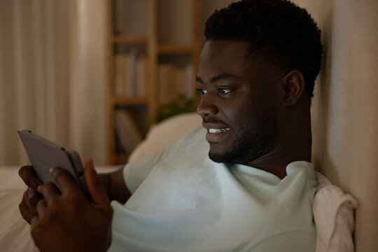 African American adult male reclining on bed at home holding tablet with protective case, smiling