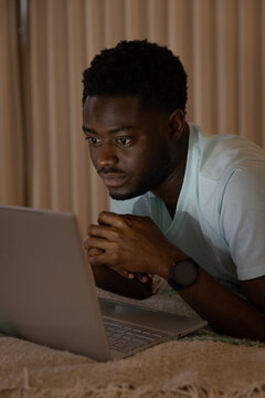 African American man propping on elbows on bed, wearing blue shirt and smartwatch viewing laptop